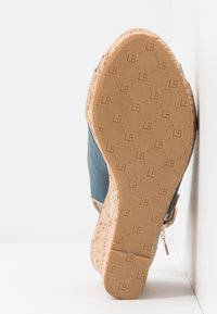 Chaussure compensée avec une semelle en liège, dessus en tissu bleu, accentuée de matière beige, présentant une texture lisse et un logo embossé sur la semelle.