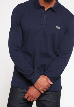 Mann trägt ein marineblaues Langarm-Poloshirt mit grünem Krokodil-Logo auf der Brust und dunkle Jeans, Hände vor dem Körper verschränkt.