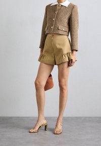 Vrouw in een bruin houndstooth cropped jack met witte kraag, khaki shorts met volants, gouden strappy hakken en een kleine bruine handtas.
