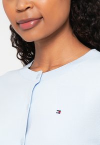 Hele sinine kardigan ümmarguse kaelusega, millel on nööbid ja rinnal väike tikandpunane, valge ja sinine logo.