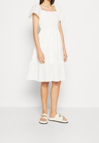 Robe blanche, longueur genoux, avec un décolleté carré, de courtes manches bouffantes et une jupe à volants. Portée avec des sandales à glissière beiges avec des accessoires décoratifs.