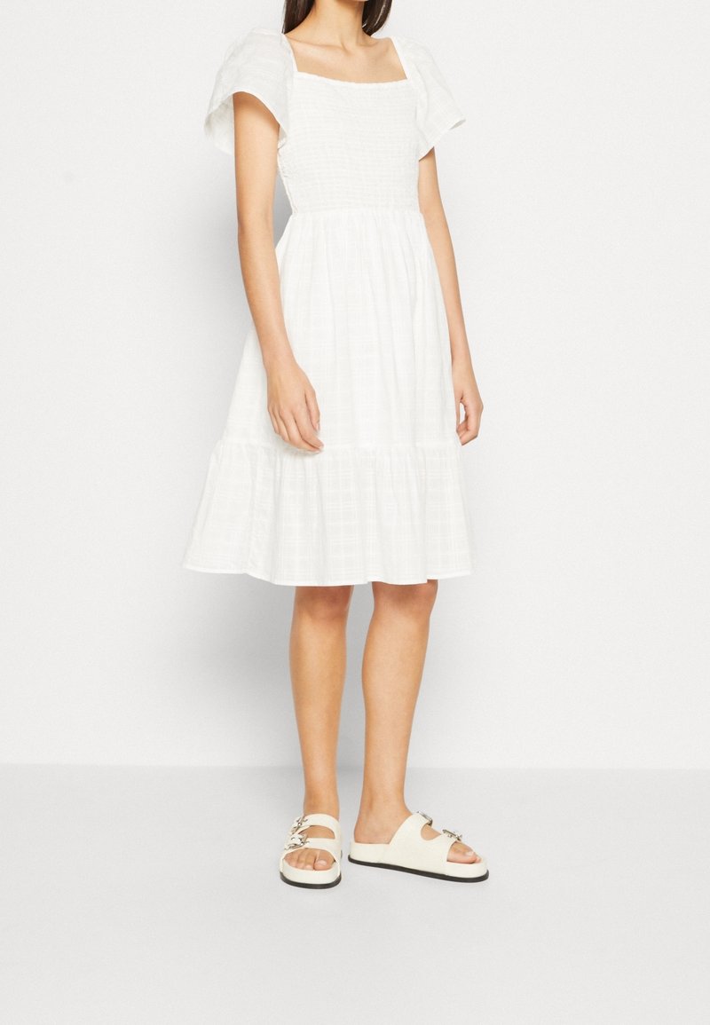 Robe blanche, longueur genoux, avec un décolleté carré, de courtes manches bouffantes et une jupe à volants. Portée avec des sandales à glissière beiges avec des accessoires décoratifs.