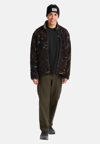 Schwarze und braune gemusterte Fleecejacke mit Reißverschluss, getragen über einem schwarzen Shirt und kombiniert mit olivgrünen Cargo-Hosen und grauen Sneakers.