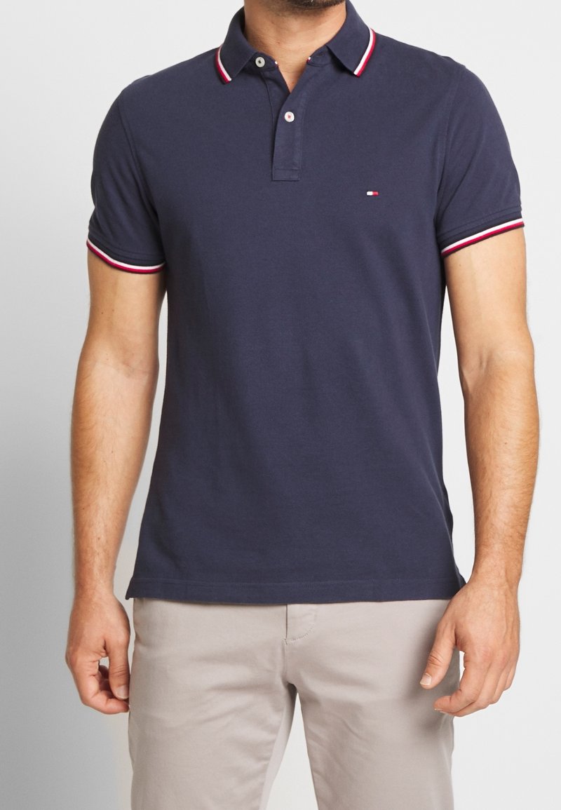 Homme portant un polo bleu marine avec des bordures rouges et blanches sur le col et les manches, associé à un pantalon beige.