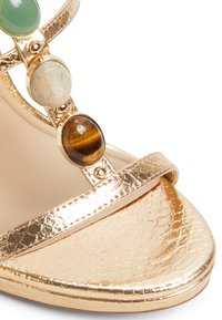 Gouden metallic hoge hak sandaal met een gestructureerde afwerking, voorzien van drie cirkelvormige steenaccenten in groen, beige en bruin. Bandjesontwerp.