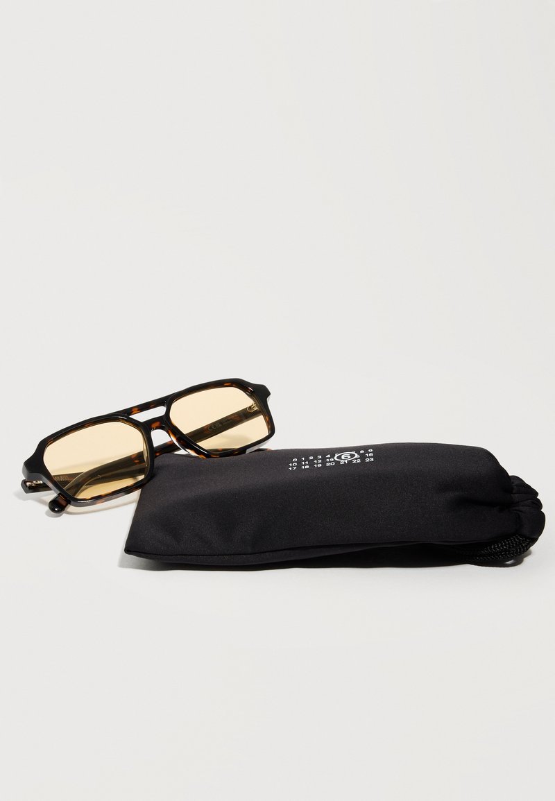 Lunettes de soleil rectangulaires écaille de tortue avec des verres jaunes reposant sur une pochette noire souple avec des chiffres blancs imprimés.