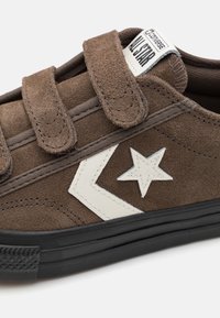 Zapato de ante marrón con correas de velcro, que presenta un diseño de estrella blanca y chevron. Suela de goma negra con agarre texturizado.