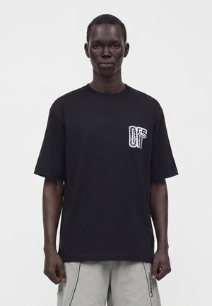 Άνδρας που φοράει μαύρο φαρδύ T-shirt με μικρό λευκό σχέδιο στο στήθος και ανοιχτό γκρι σορτς σε απλό φόντο.