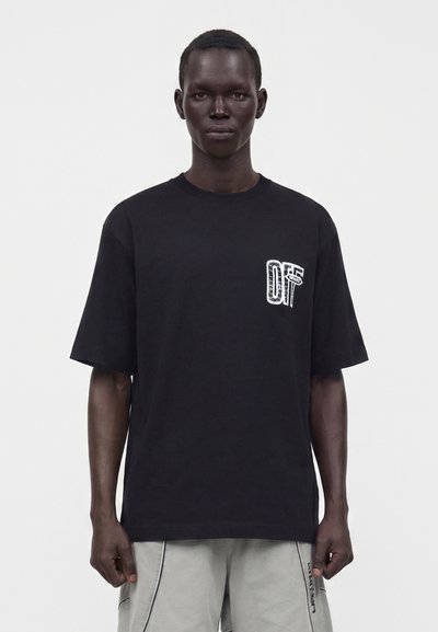 Άνδρας που φοράει μαύρο φαρδύ T-shirt με μικρό λευκό σχέδιο στο στήθος και ανοιχτό γκρι σορτς σε απλό φόντο.