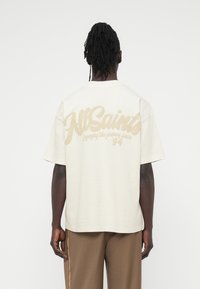 Φαρδιά κοντομάνικη μπλούζα σε ανοιχτό μπεζ χρώμα από βαμβάκι, με μεγάλο λευκό λογότυπο στην πλάτη, κοντά μανίκια και άνετη εφαρμογή.