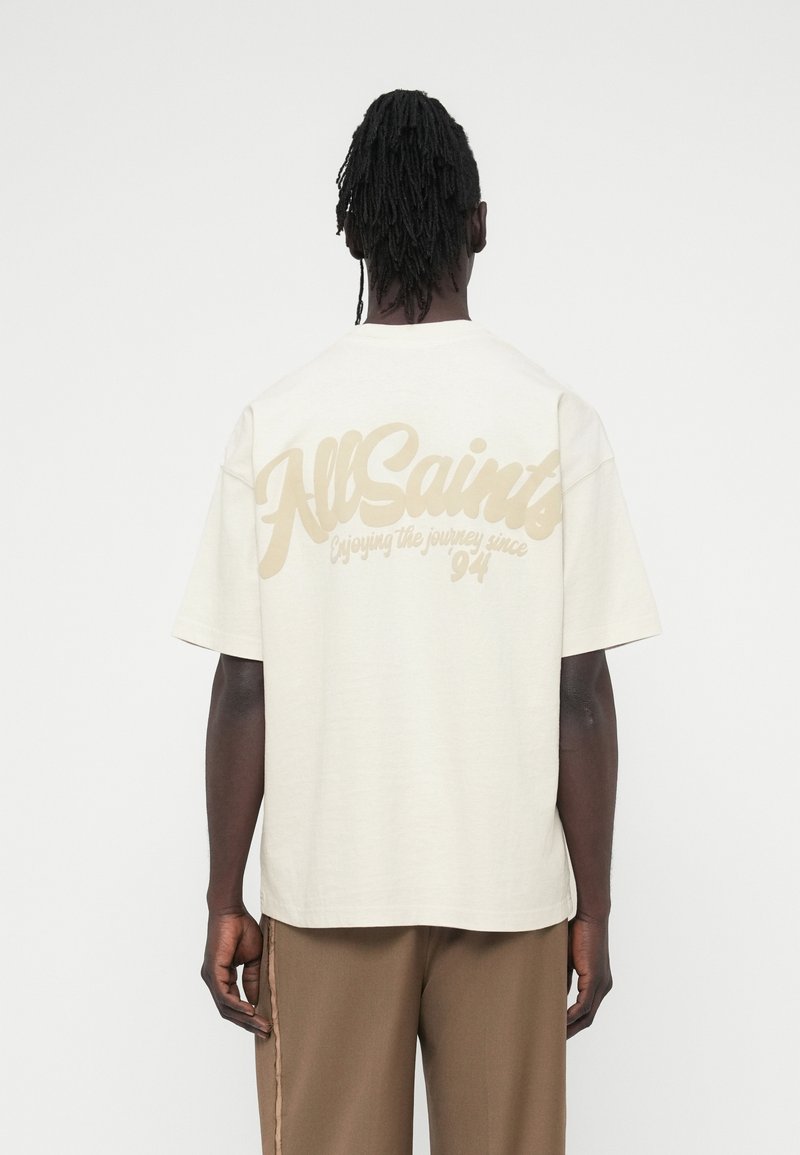 Φαρδιά κοντομάνικη μπλούζα σε ανοιχτό μπεζ χρώμα από βαμβάκι, με μεγάλο λευκό λογότυπο στην πλάτη, κοντά μανίκια και άνετη εφαρμογή.