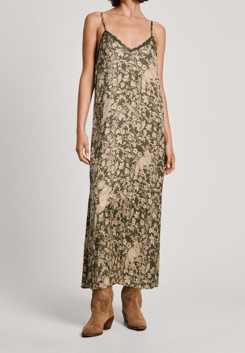 Femme portant une robe maxi sans manches vert olive avec un motif beige floral et hibou, associée à des bottines en suède marron clair.