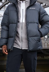 Czarna puchowa kurtka z kapturem i zamkiem, z szarym polarowym podkładem i ciemnymi spodniami, odpowiednia na zimną pogodę.