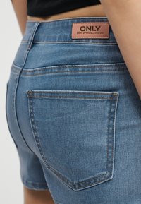 Κοντινό πλάνο μπλε τζιν σορτς με δερμάτινο σήμα μάρκας στην πίσω ζώνη, με την ετικέτα «ONLY Blue Denim anno 1995».