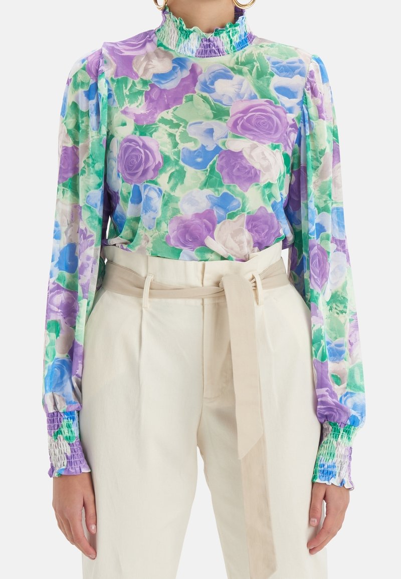 Femme portant un chemisier floral à col montant en violet, vert et bleu, rentré dans un pantalon crème taille haute avec une ceinture assortie.
