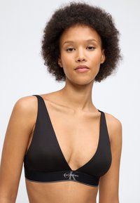 Νεαρή γυναίκα με σγουρά μαλλιά που φοράει ένα μαύρο bralette Calvin Klein με βαθύ V ντεκολτέ σε απλό φόντο.