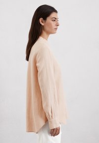 Licht perzikkleurige blouse met lange mouwen, een ronde kraag, rechte zoom en knoopmanchetten; gemaakt van zachte, lichte stof.