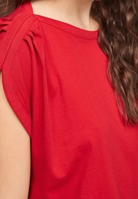 Top rosso a maniche corte realizzato in morbido cotone. Caratterizzato da dettagli arricciati sulla spalla, scollatura arrotondata e texture liscia per garantire comfort.
