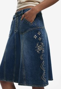 Gonna in denim con forma svasata, di colore blu scuro, con due tasche frontali e ricami floreali decorativi sul lato sinistro.