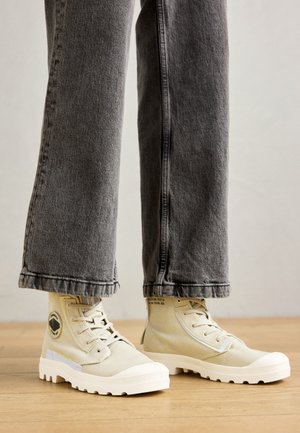 Light beige high-top boots made of canvas with rubber soles, rounded toe caps, and contrasting white accents. Worn with gray wide-leg jeans.
