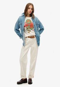 Camisa de mezclilla, camiseta gráfica crema con diseño rojo y verde, pantalones beige y mocasines marrón oscuro, posando con las manos en las caderas.