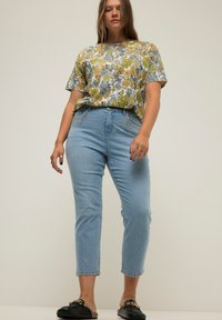 Top floreale a maniche corte giallo, verde e blu; jeans a vita alta in lavaggio chiaro; scarpe slip-on nere con accenti dorati.