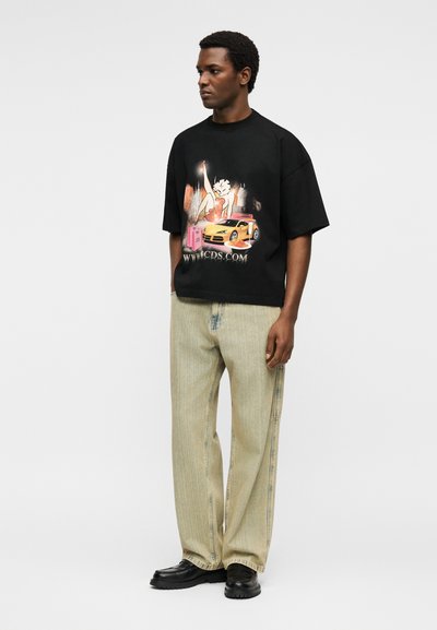 Άνδρας που φοράει μαύρο φαρδύ T-shirt με εικόνα μιας καρτούν γυναίκας, κίτρινο σπορ αυτοκίνητο και κείμενο "WWW.CDS.COM", μπεζ παντελόνι και μαύρα παπούτσια.