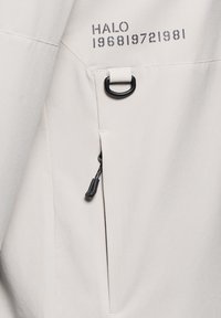 Jasnoszara kurtka o gładkiej teksturze, z odsłoniętym czarnym zamkiem i detalem w formie pętli. Nadrukowany tekst brzmi "HALO 196819721981."