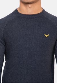 Primer plano de una persona que lleva un suéter de canalé en azul marino oscuro con un pequeño logo amarillo bordado en el lado izquierdo del pecho.