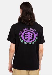 T-shirt en coton noir avec un grand motif circulaire au dos en violet et vert, affichant les mots "Terre, Eau, Feu, Air" avec un symbole d'arbre.