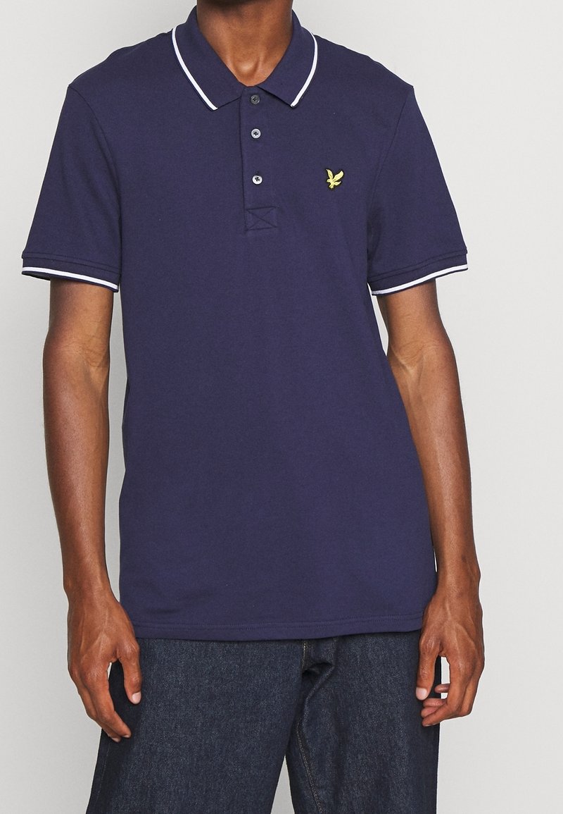 Polo shirt blu navy con colletto e bordi delle maniche bianchi, dotato di una patta con tre bottoni e logo ricamato sul lato sinistro del petto. Tessuto in cotone.