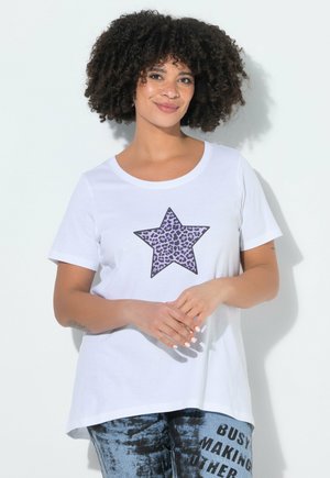 Frau mit lockigem Haar, die ein weißes T-Shirt mit einem lila Leopardstern und blauen Batikhosen mit schwarzem Text trägt.