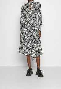Černo-bílé vzorované šaty s vysokým límcem, detaily ve tvaru klíčové dírky, dlouhými rukávy a asymetrickým lemem, doplněné černými platformovými botami.