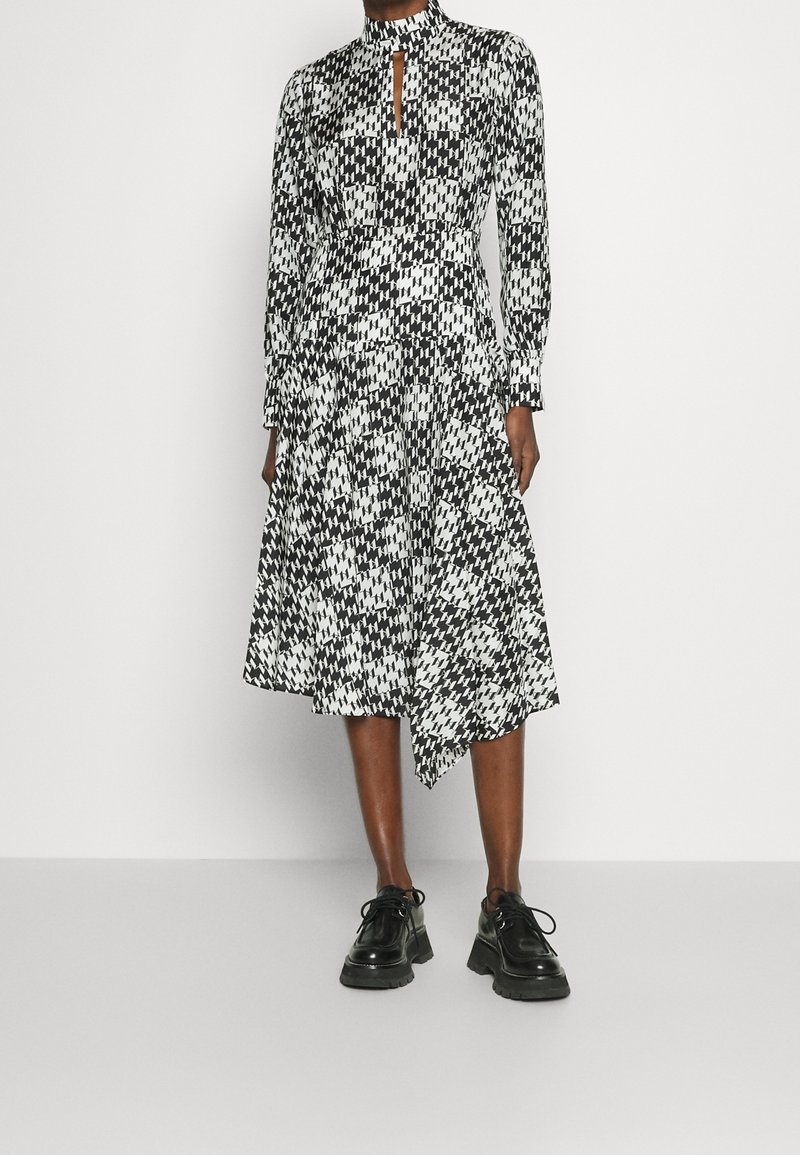 Černo-bílé vzorované šaty s vysokým límcem, detaily ve tvaru klíčové dírky, dlouhými rukávy a asymetrickým lemem, doplněné černými platformovými botami.