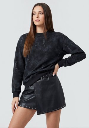 Donna con capelli lunghi e lisci che indossa una felpa nera marmorizzata e una minigonna in pelle nera con borchie metalliche e cintura, posando con la mano sul fianco.