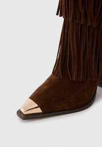 Bota de ante marrón con detalles de flecos largos. Presenta una punta afilada con una punta metálica dorada y una suela de goma plana.