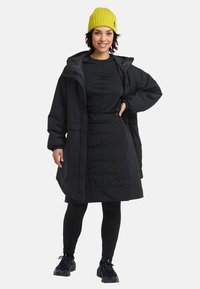Zwarte gewatteerde jas met een capuchon, gecombineerd met een zwart top en rok, zwarte legging en een felgele gebreide muts.