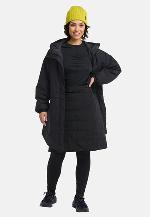 Zwarte gewatteerde jas met een capuchon, gecombineerd met een zwart top en rok, zwarte legging en een felgele gebreide muts.