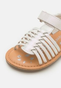 Sandalia infantil de piel blanca con parte superior trenzada, correa ajustable y suela beige con estampado floral plateado en la plantilla.