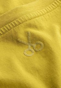 Geel katoenen stof met een zachte textuur, voorzien van een subtiele schaarafbeelding in een lichtere tint. De rand heeft een geribbelde afwerking.