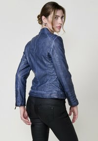 Giacca di pelle blu con un design aderente, superficie texturizzata e dettagli di cucitura diagonale. Cerniere sui polsini e colletto strutturato.