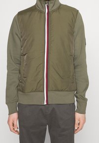 Kurtka na zamek w oliwkowozielonym kolorze z wysokim kołnierzem, miękką fakturą, czerwono-biało paskowanym zamkiem błyskawicznym i bocznymi kieszeniami. Styl dziecięcy.