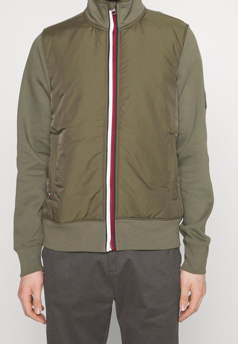 Kurtka na zamek w oliwkowozielonym kolorze z wysokim kołnierzem, miękką fakturą, czerwono-biało paskowanym zamkiem błyskawicznym i bocznymi kieszeniami. Styl dziecięcy.