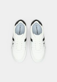 Λευκά αθλητικά παπούτσια με μαύρες λεπτομέρειες. Λείο συνθετικό επάνω μέρος, σχέδιο με κορδόνια, επενδυμένο κολάρο και λογότυπο στη πλευρά. Μινιμαλιστικό στυλ.