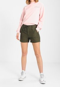 Light pink sweatshirt with "Good Girl" text in white, paired with olive green cargo shorts. White high-top sneakers complete the outfit.