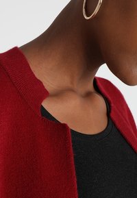 Červený vlněný cardigan s kulatým výstřihem, strukturální pletení a otevřeným předním dílem, nošený přes černý top, který zvýrazňuje kontrast mezi látkou a barvou oděvu.