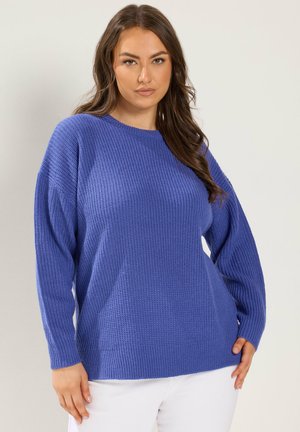 Vrouw met lang bruin haar die een gestructureerde blauwe trui en witte broek draagt, staand tegen een effen lichte achtergrond.