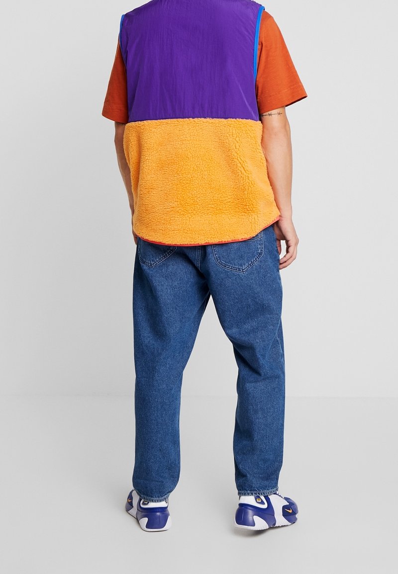 Gilet in pile colorato viola, arancione e blu su una maglietta arancione, abbinato a jeans blu larghi e sneakers bianche con accenti viola.