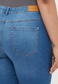 Mėlynos džinsinės kelnės su vidutiniu juosmens pakėlimu, turinčios šviesiai rudos odos etiketę, nugaros kišenės detales ir tradicinį siuvinėjimą.