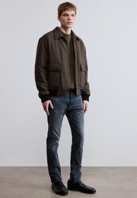 Brun jakke i ullblanding med krage, to frontlommer, ribbestrikkede ermepartier, kombinert med grå jeans og svarte sko.